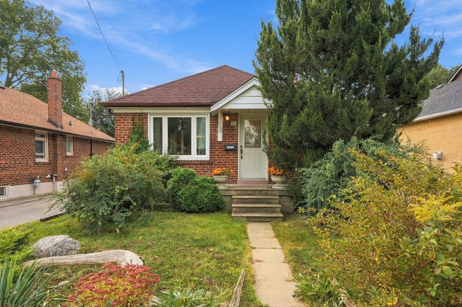 Front of the red-brick bungalow with mature evergreens and a stone path to the front steps